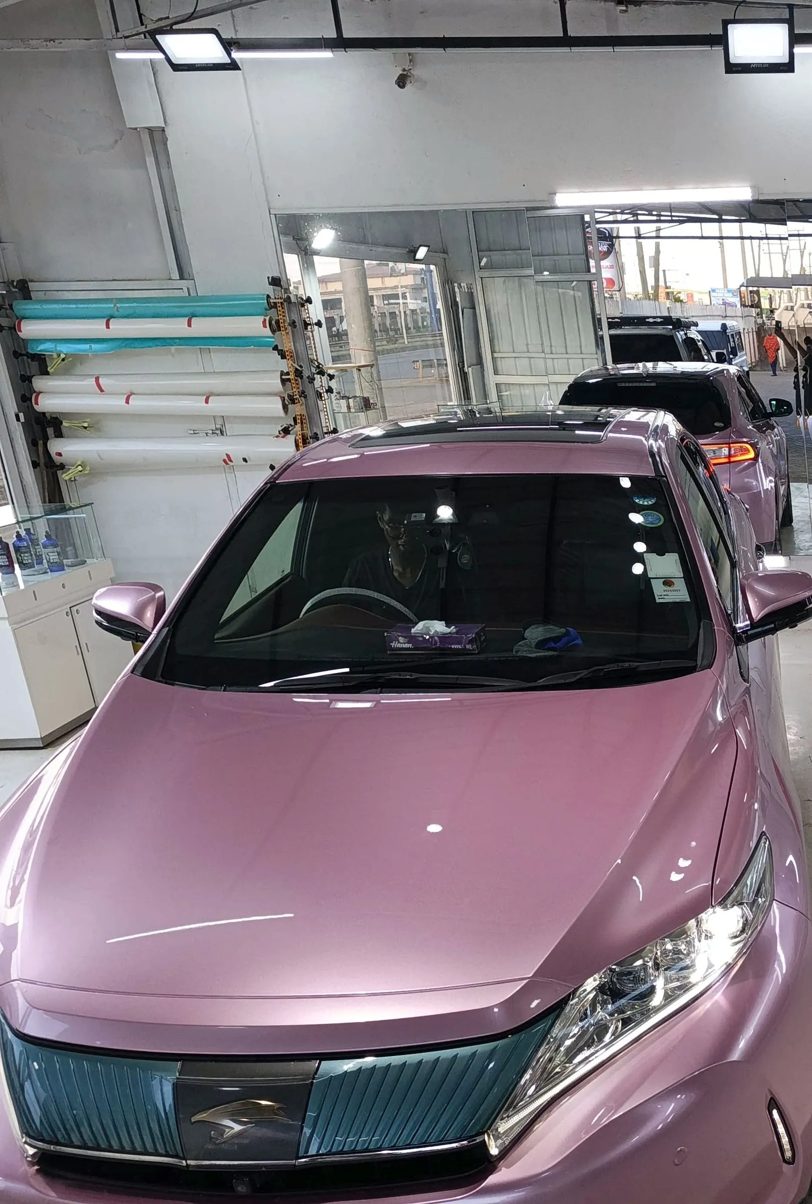 Front view of a Toyota Harrier wrapped in soft lavender, with rolls of vinyl on the workshop wall behind